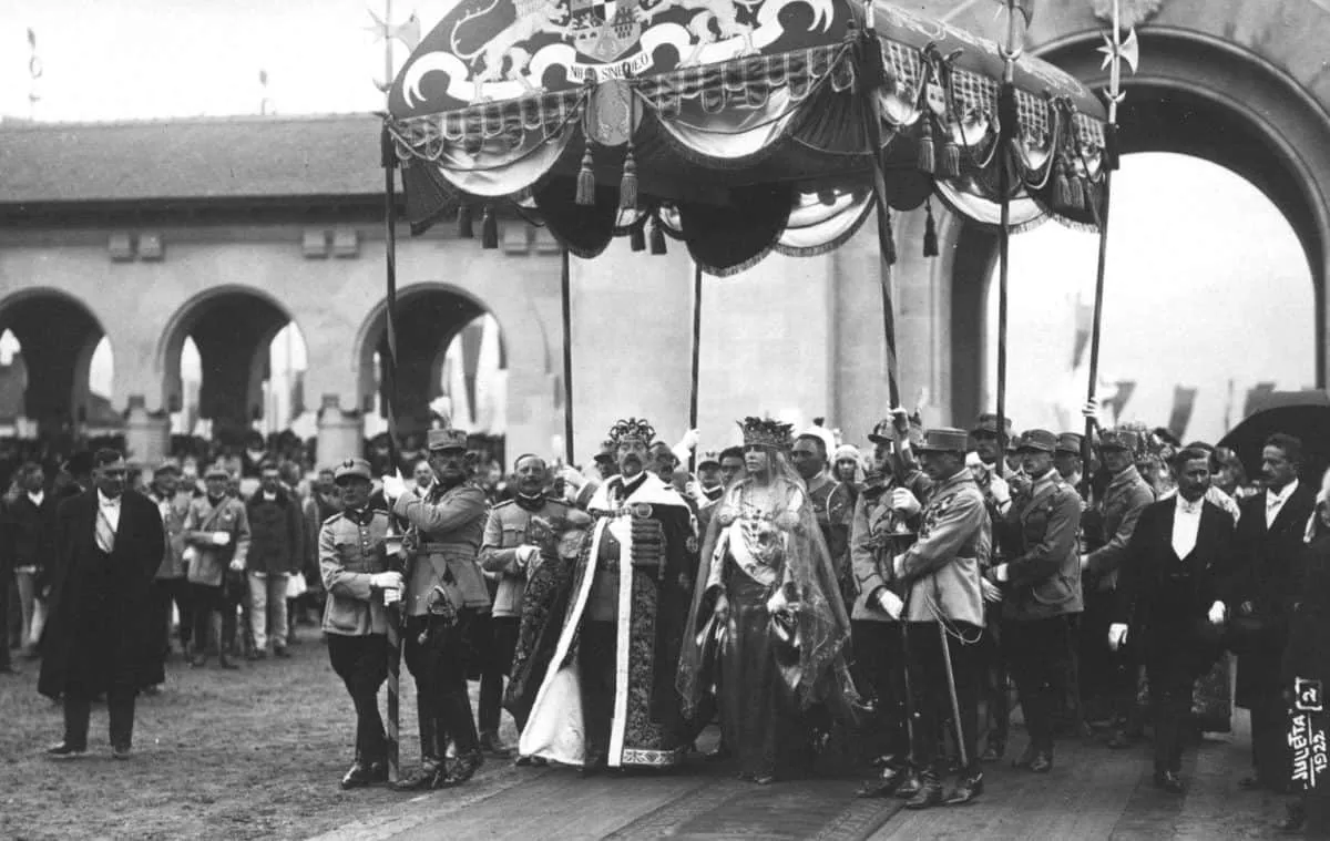 Sub un baldachin ornamentat stau două persoane cu veșminte lungi, înconjurate de bărbați în uniforme și costume; în fundal se văd arcade.