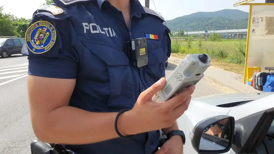 Un polițist în uniformă ține un aparat electronic lângă o mașină, pe marginea unui drum.