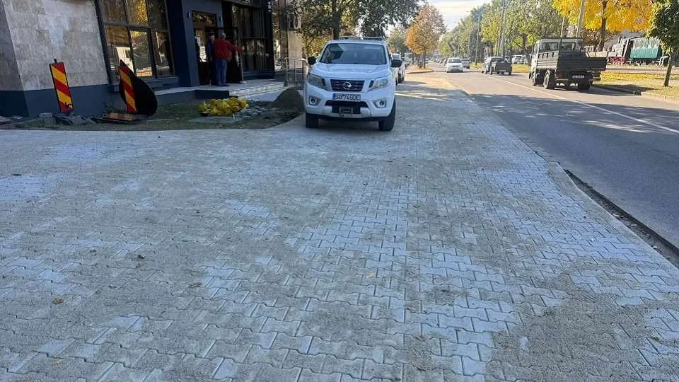 Un trotuar lat, pavat cu dale gri, ocupă prim-planul; o camionetă albă este parcată lângă o clădire, iar carosabilul se află în dreapta.