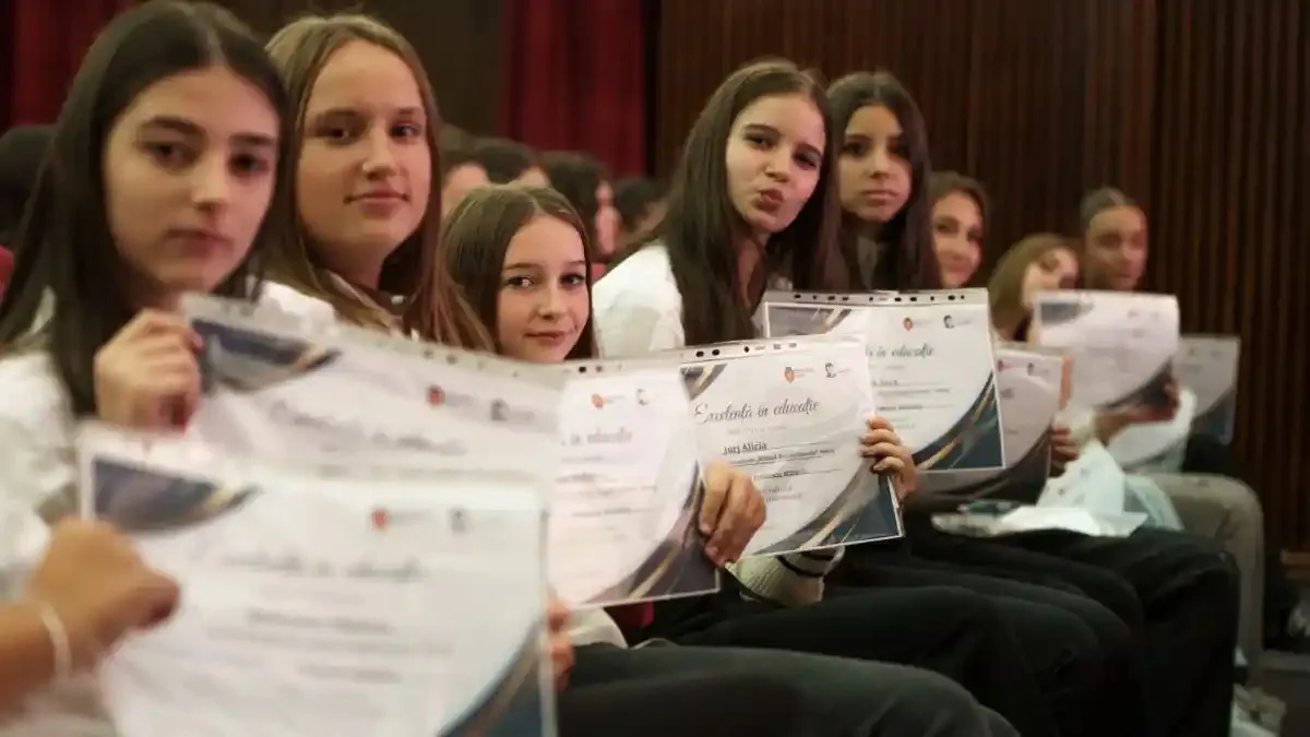 Mai multe tinere stau așezate pe scaune, într-o sală cu draperii roșii, și țin diplome tipărite.