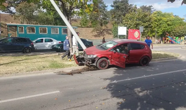 O mașină roșie avariată este oprită lângă un stâlp înclinat; câteva persoane și alte mașini sunt vizibile în apropiere.