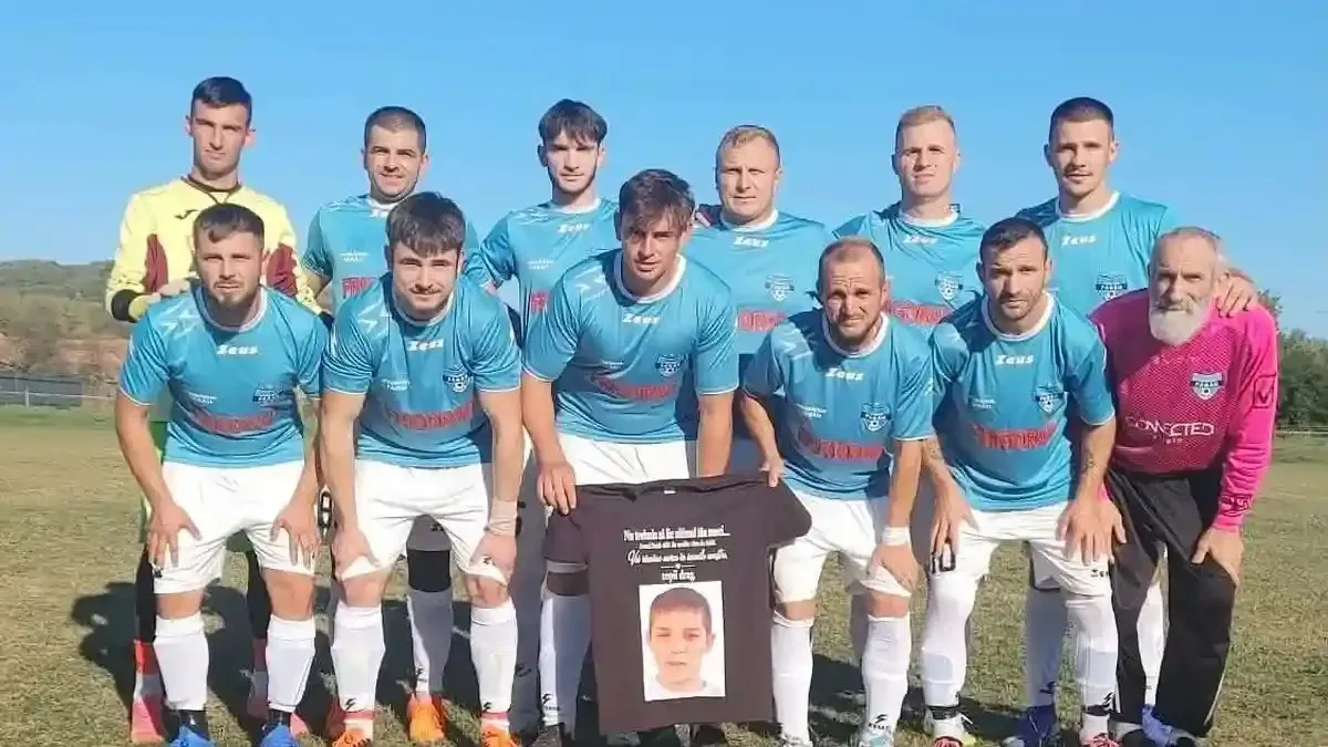 Unsprezece bărbați în echipamente sportive albastre și albe pozează pe un teren, ținând un tricou negru imprimat cu fotografia unui copil.