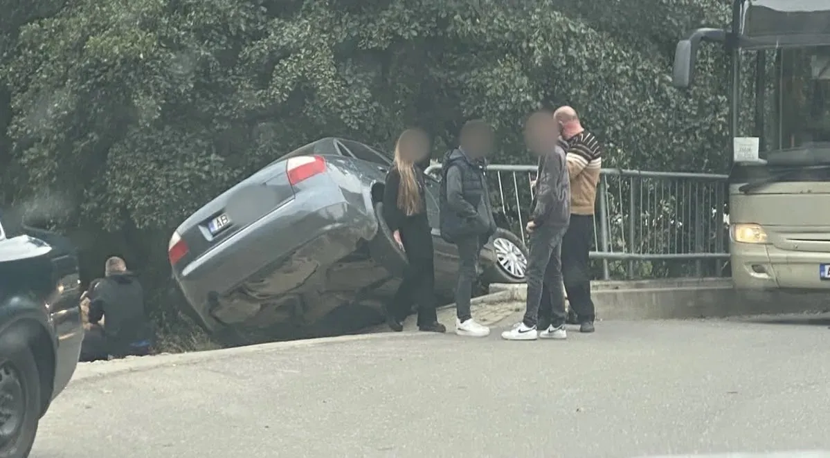 Un autoturism gri este înclinat lângă un parapet; mai multe persoane stau în apropiere, iar un autobuz se vede în dreapta.