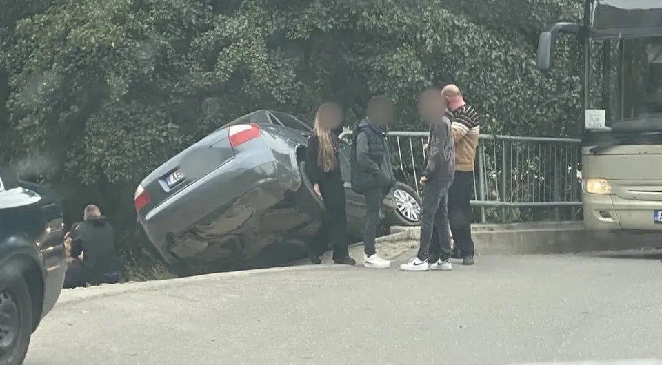 Un autoturism gri este înclinat lângă un parapet; mai multe persoane stau în apropiere, iar un autobuz se vede în dreapta.