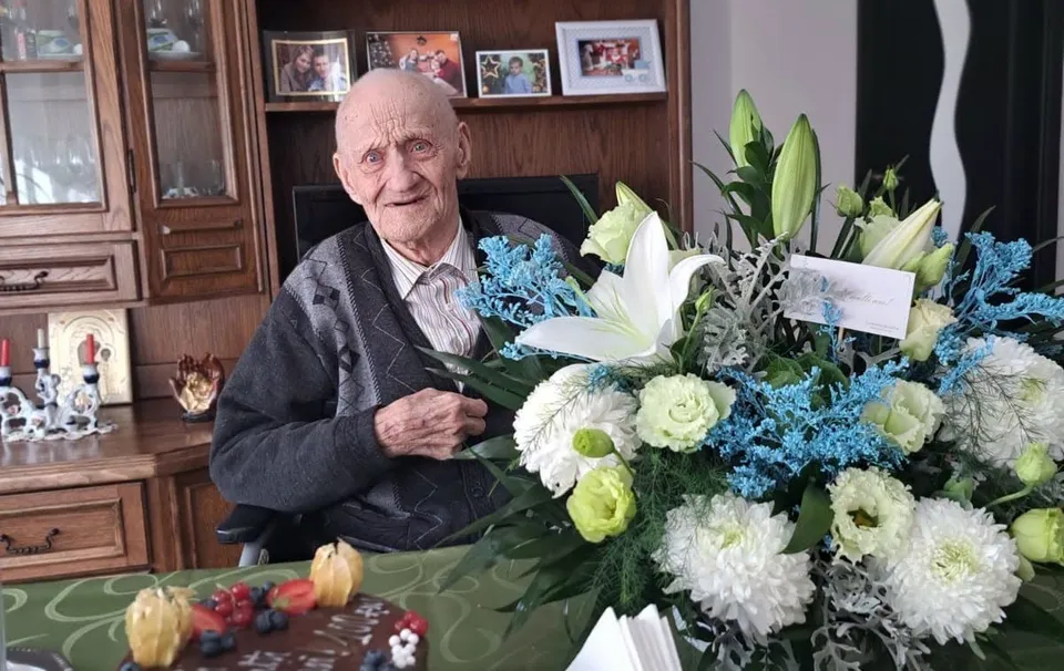 Un bărbat în vârstă stă lângă un buchet mare de flori, cu un tort decorat aflat în prim-plan.