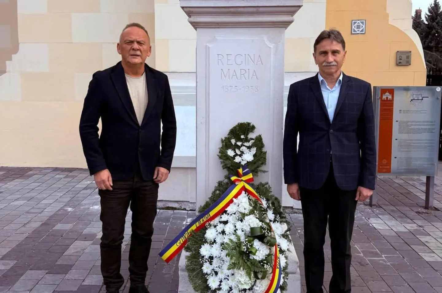 Doi bărbați în sacouri închise la culoare stau de o parte și de alta a unui soclu inscripționat, lângă coroane de flori albe cu panglici tricolore.