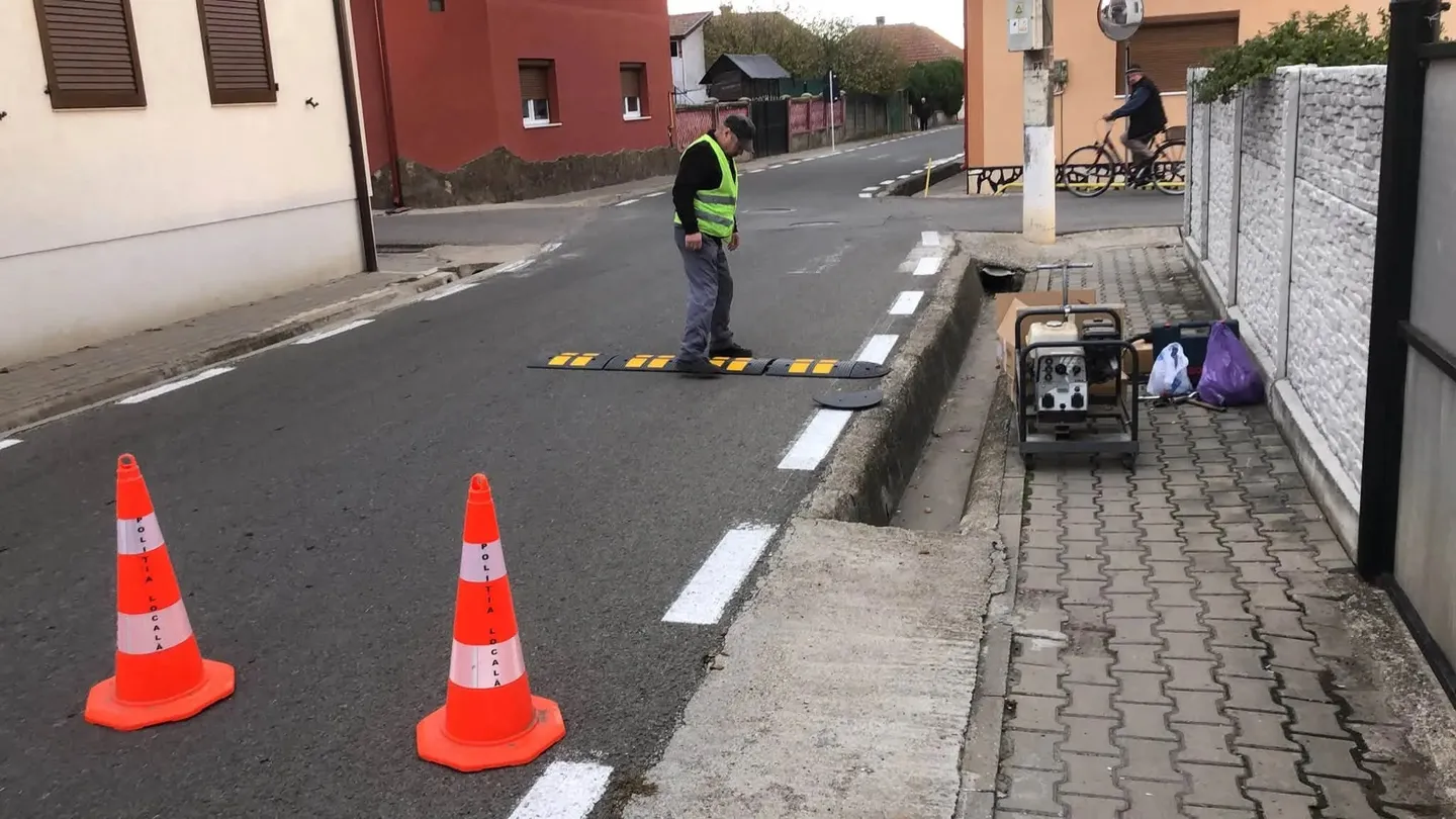 Pe o stradă, un muncitor cu vestă reflectorizantă stă lângă un limitator de viteză; două conuri portocalii sunt în față, iar un utilaj este pe trotuar.
