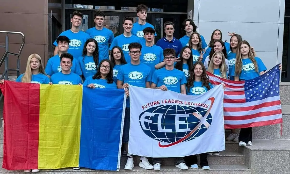 Un grup de tineri în tricouri albastre stă pe trepte, în spatele steagurilor României și SUA și al unui banner FLEX.