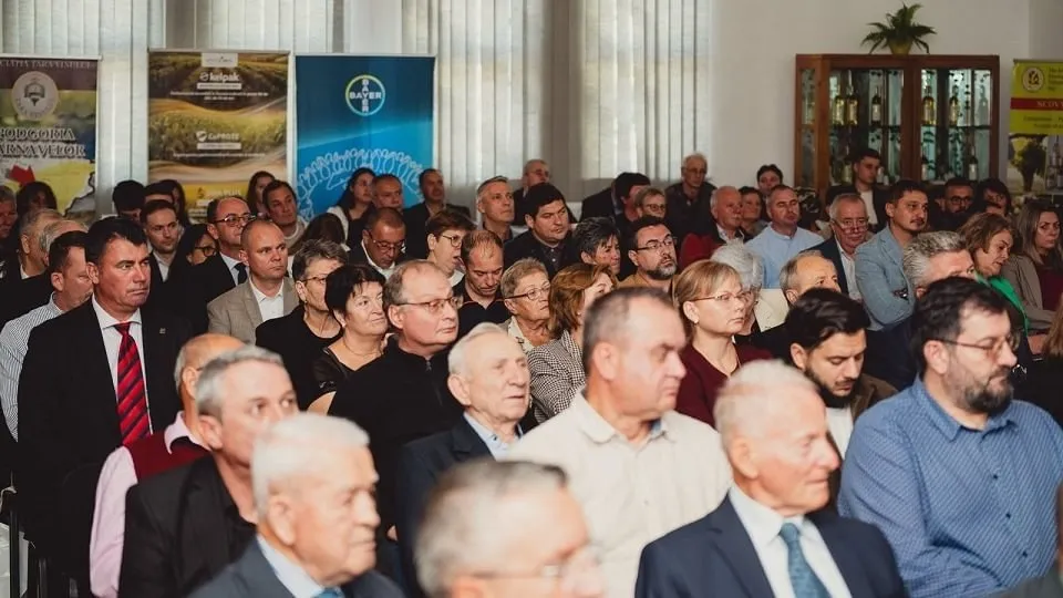 Mai multe persoane, îmbrăcate predominant în haine elegante, stau așezate în rânduri într-o încăpere, cu bannere vizibile pe peretele din spate.