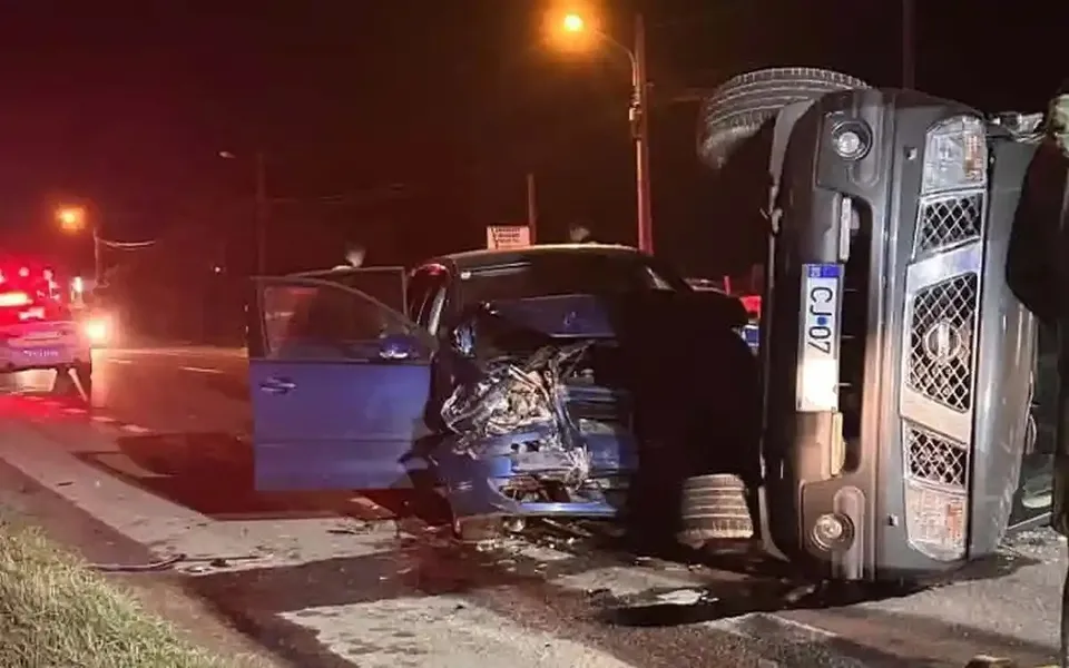 Noaptea, un autoturism albastru cu portiera deschisă și partea din față avariată se află lângă un vehicul răsturnat pe o parte.