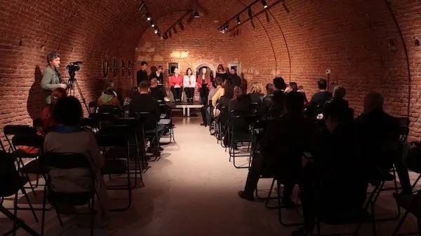 Într-o sală cu pereți și tavan boltit din cărămidă, persoane stau pe scaune orientate spre un grup aflat lângă lucrări expuse pe perete.
