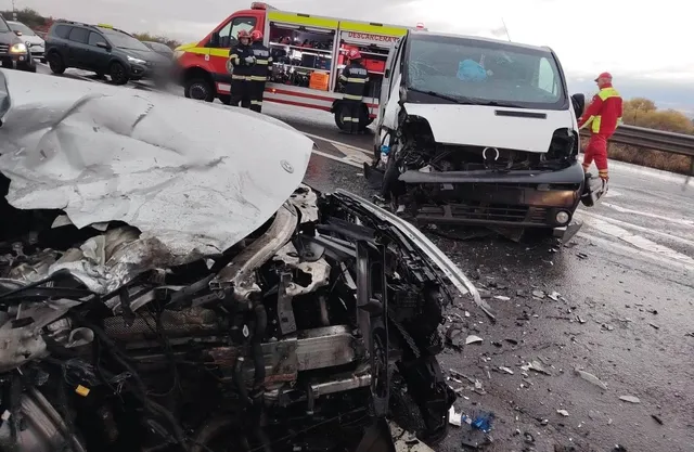 În prim-plan se vede un autoturism grav avariat, iar în spate se află o autoutilitară avariată, o autospecială și o persoană în haine roșii.