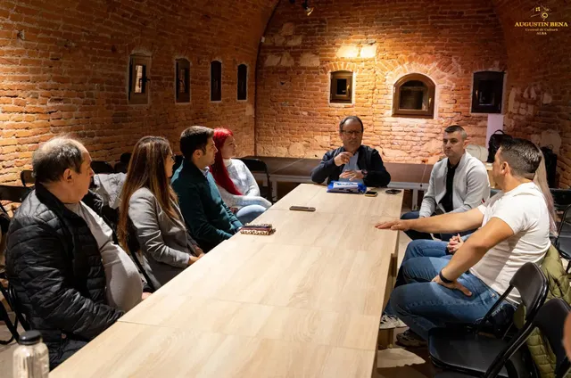 Mai multe persoane stau pe scaune în jurul unei mese lungi, într-o încăpere cu pereți de cărămidă; pe masă se află obiecte mici.