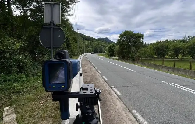 Un aparat albastru montat pe un trepied se află lângă un drum asfaltat, cu indicatoare, copaci și dealuri în fundal.