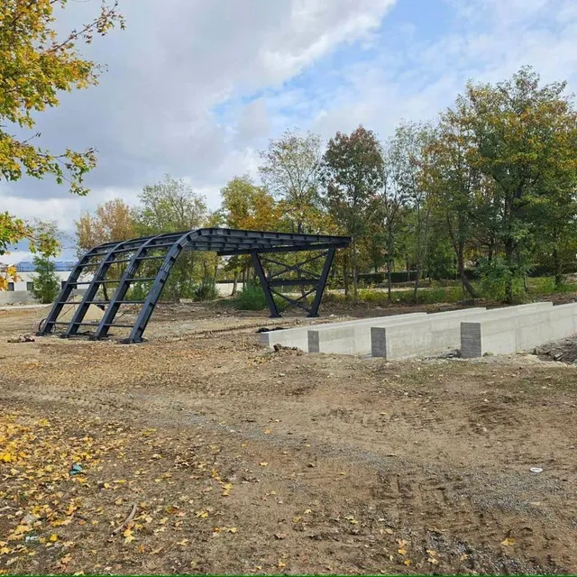 Pe un teren de pământ și pietriș se află o structură metalică neagră arcuită și o platformă joasă din beton, cu copaci în spate.