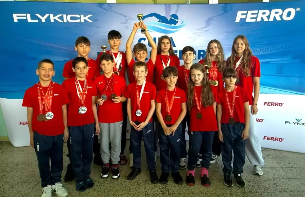 Un grup de copii și adolescenți în tricouri roșii poartă medalii, iar câțiva țin cupe, în fața unui panou albastru.