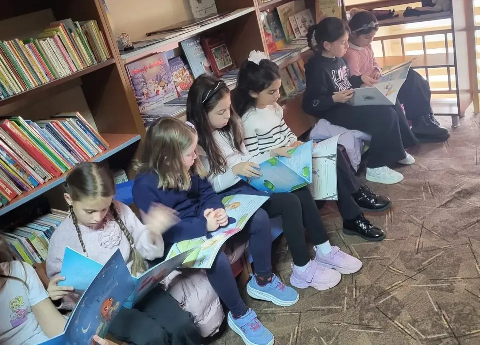 Mai multe fete stau pe podea și răsfoiesc cărți deschise, lângă rafturi cu cărți; unele poartă ghiozdane.