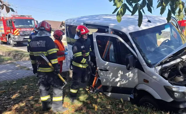 Un microbuz alb are partea frontală avariată; lângă el se află trei persoane în echipament de intervenție, iar în fundal se vede o autospecială roșie.
