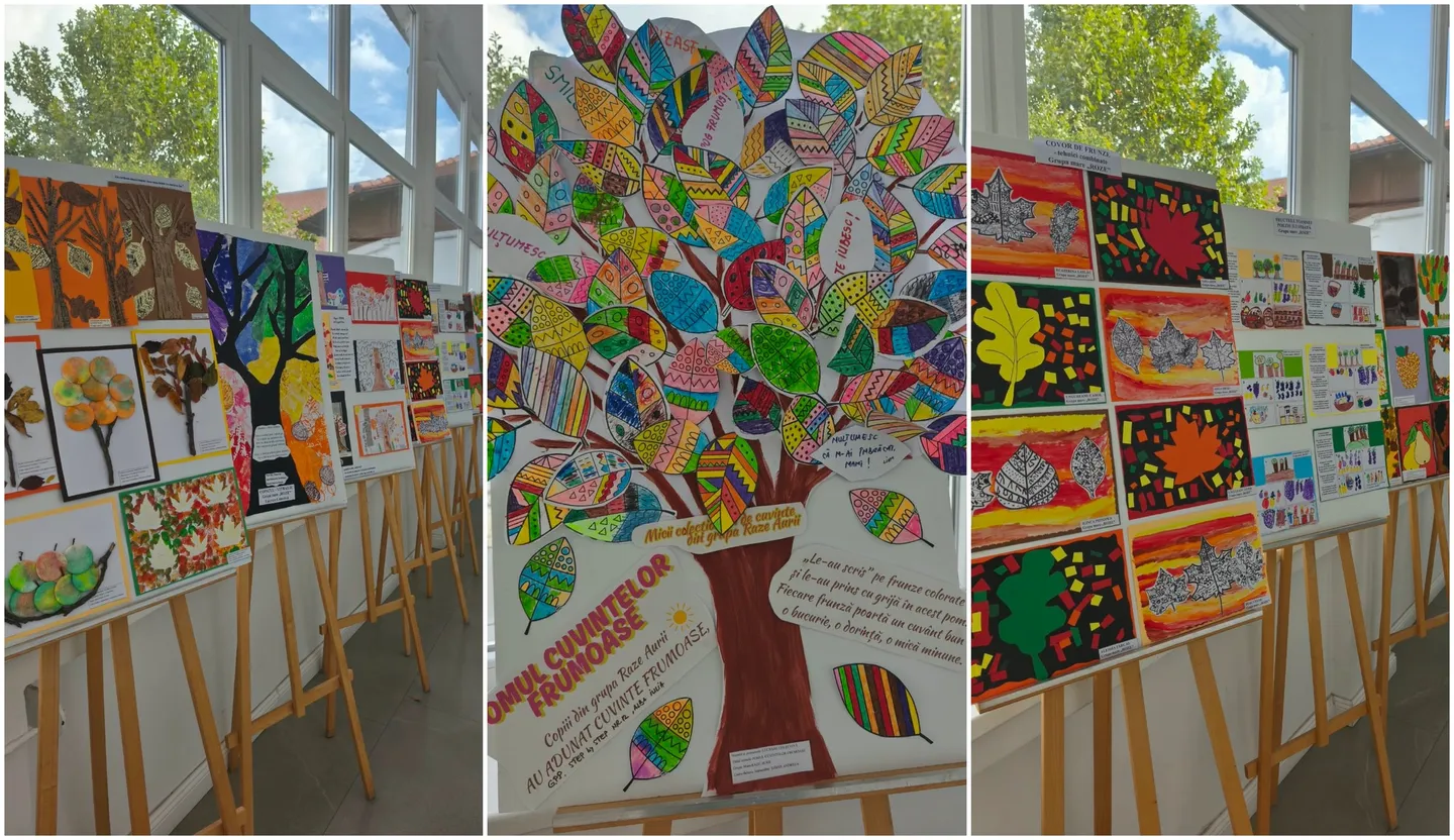Mai multe panouri cu desene colorate sunt așezate pe șevalete lângă ferestre; panoul central prezintă un copac cu frunze multicolore.