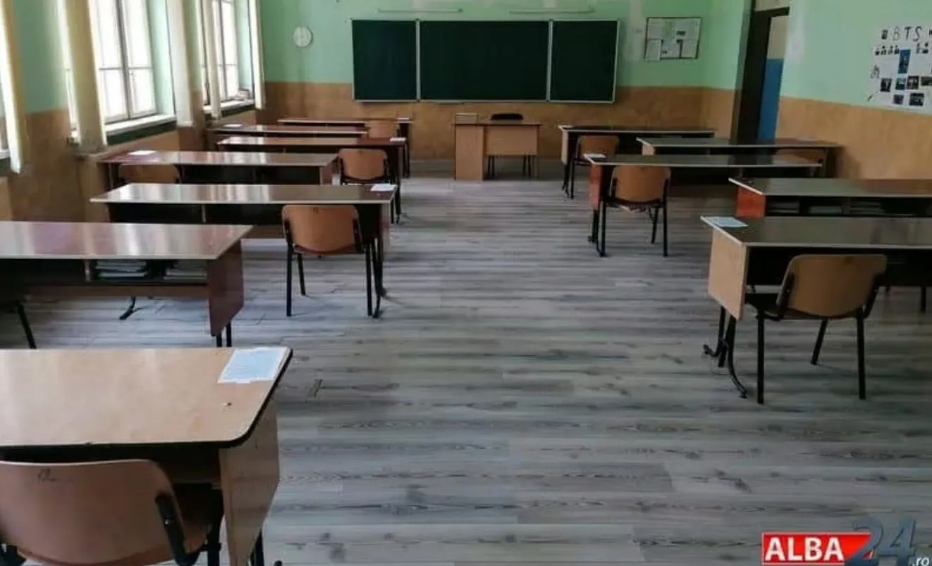 O sală goală are bănci și scaune dispuse în rânduri, o tablă pe peretele din spate și ferestre pe peretele din stânga.
