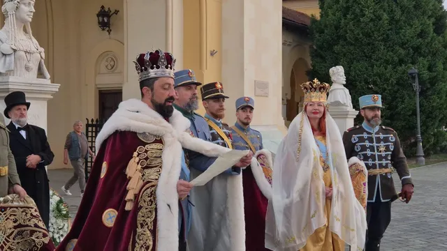 În fața unei clădiri, un bărbat cu coroană și mantie vișinie ține o foaie, lângă o femeie în veșminte deschise și alte persoane costumate.