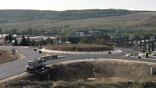 Un utilaj cu braț articulat se află lângă un drum asfaltat curbat; în prim-plan se văd pământ excavat și blocuri de beton.