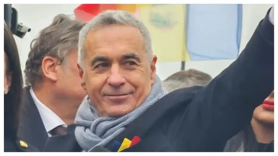 Un bărbat cu păr cărunt și fular gri stă între alte persoane, purtând la piept panglici tricolore, iar un braț apare ridicat.
