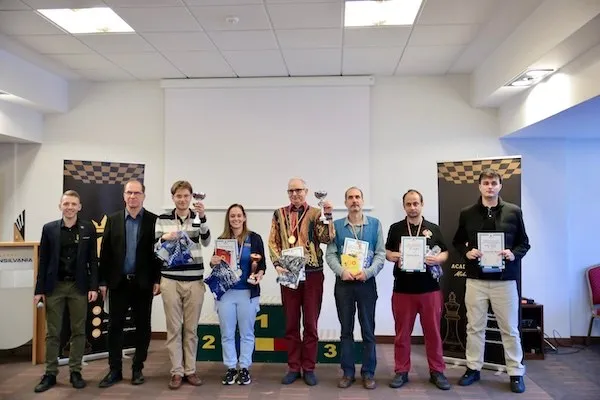Opt persoane stau în fața unui podium colorat; câteva țin diplome și obiecte mici, iar pe perete sunt două table de șah.