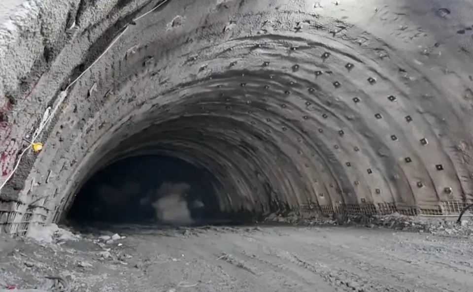 Un tunel excavat are pereți curbați cu găuri și elemente de susținere, iar pe jos se află pământ și resturi de construcție.