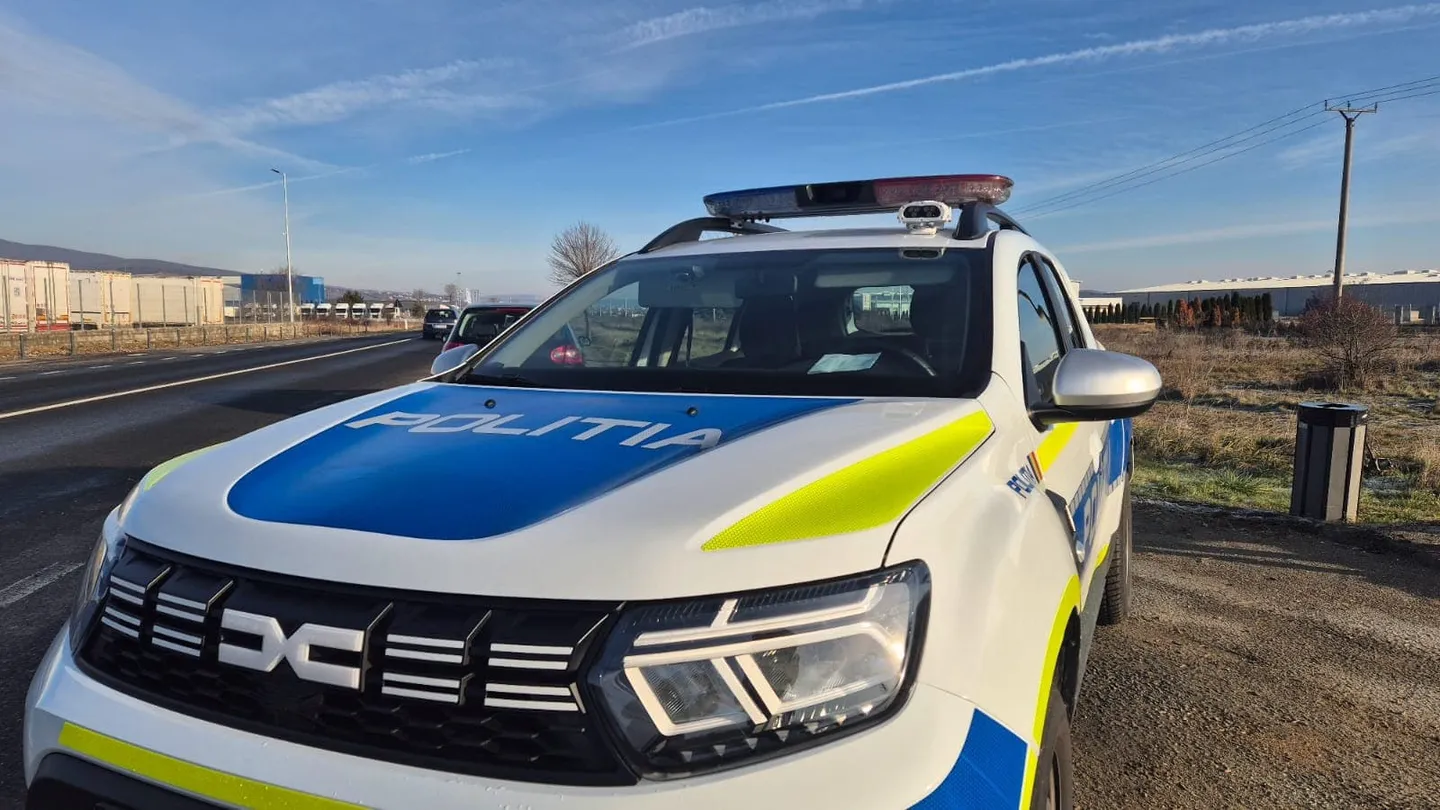Un autoturism alb de poliție, cu marcaje albastre și galbene și rampă luminoasă, este parcat lângă o șosea.