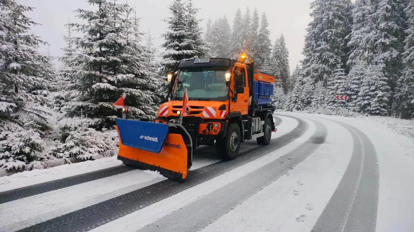 Un camion portocaliu cu lamă albastră în față se află pe un drum acoperit parțial de zăpadă, între copaci înzăpeziți.