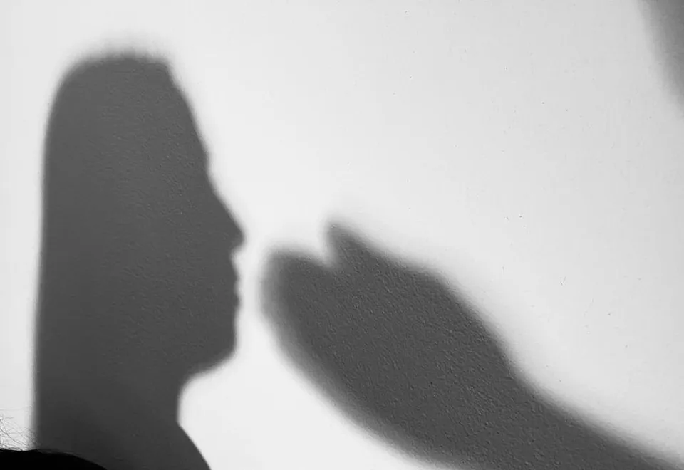 Pe un fundal deschis apar două umbre întunecate: profilul unei persoane în stânga și o mână ridicată în dreapta.