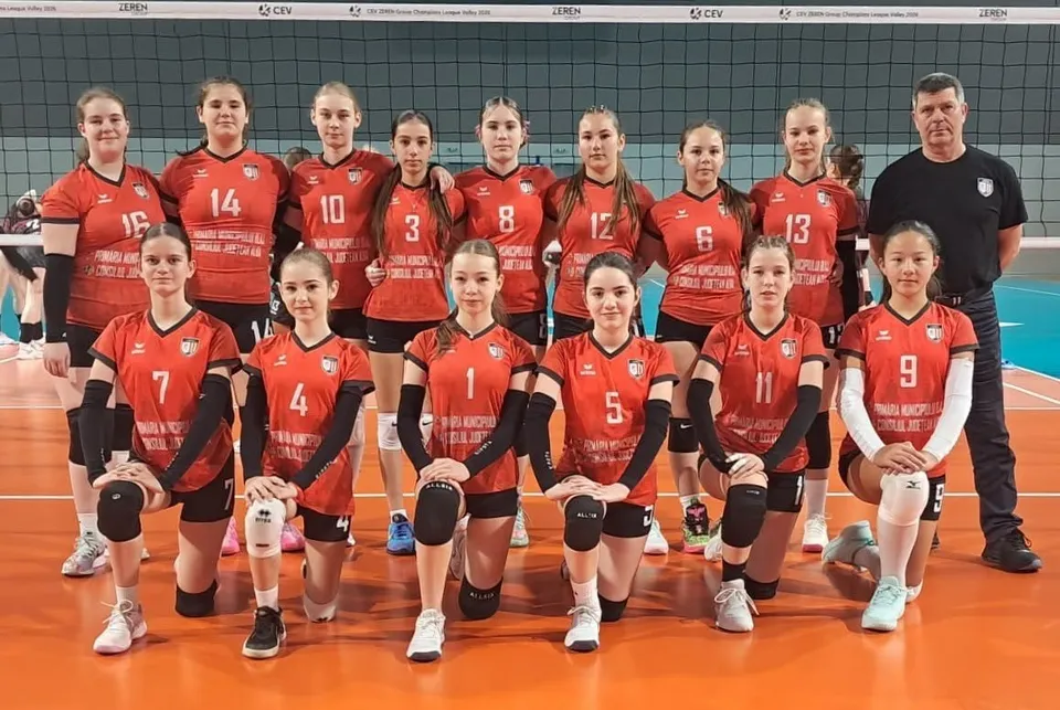 Un grup de sportive în tricouri roșii și pantaloni scurți stă în două rânduri pe o suprafață portocalie, alături de un bărbat.