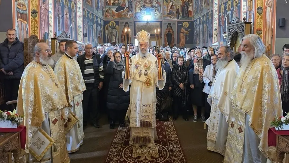 Un cleric cu veșminte aurii stă în centrul unei biserici pictate, între alți clerici, în fața unui grup de oameni.