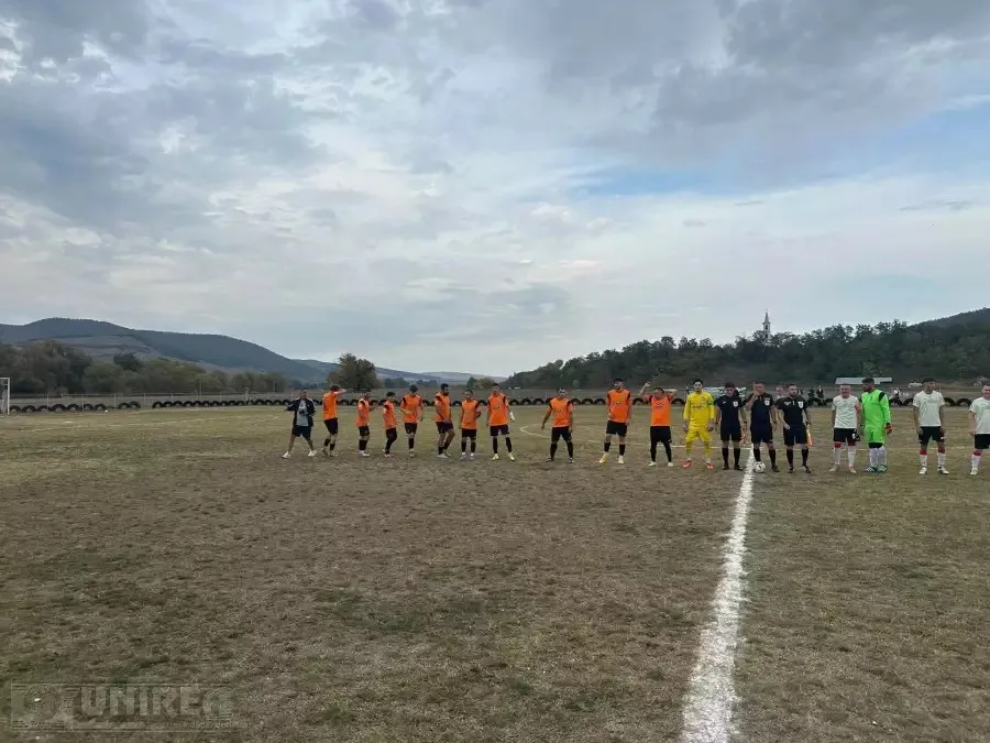 Pe un teren de fotbal, jucători în tricouri portocalii, negre și deschise la culoare stau aliniați de-a lungul liniei centrale, sub un cer înnorat.