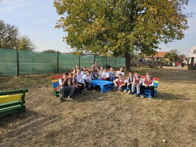 Un grup de elevi stă pe bănci viu colorate în jurul unei mese albastre, sub un copac, într-o curte împrejmuită.