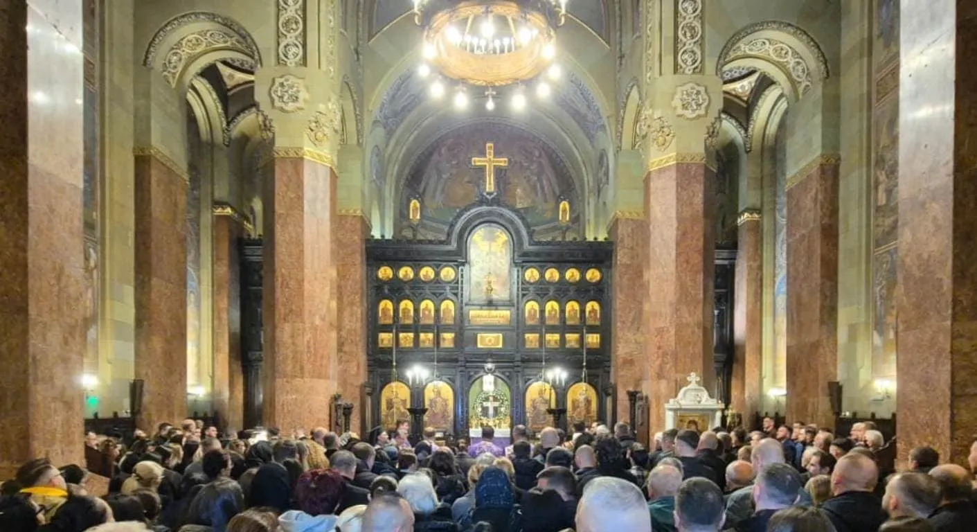 Interiorul unei catedrale prezintă o mulțime de credincioși orientați spre altarul decorat cu icoane, sub arcade, coloane și un candelabru aprins.