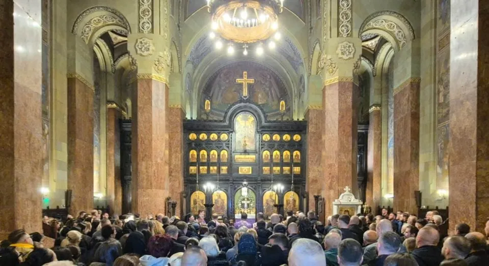 Interiorul unei catedrale prezintă o mulțime de credincioși orientați spre altarul decorat cu icoane, sub arcade, coloane și un candelabru aprins.