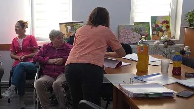 Trei femei se află lângă mese cu hârtii, dosare, pahare și sticle; una stă în picioare, iar celelalte două sunt așezate.