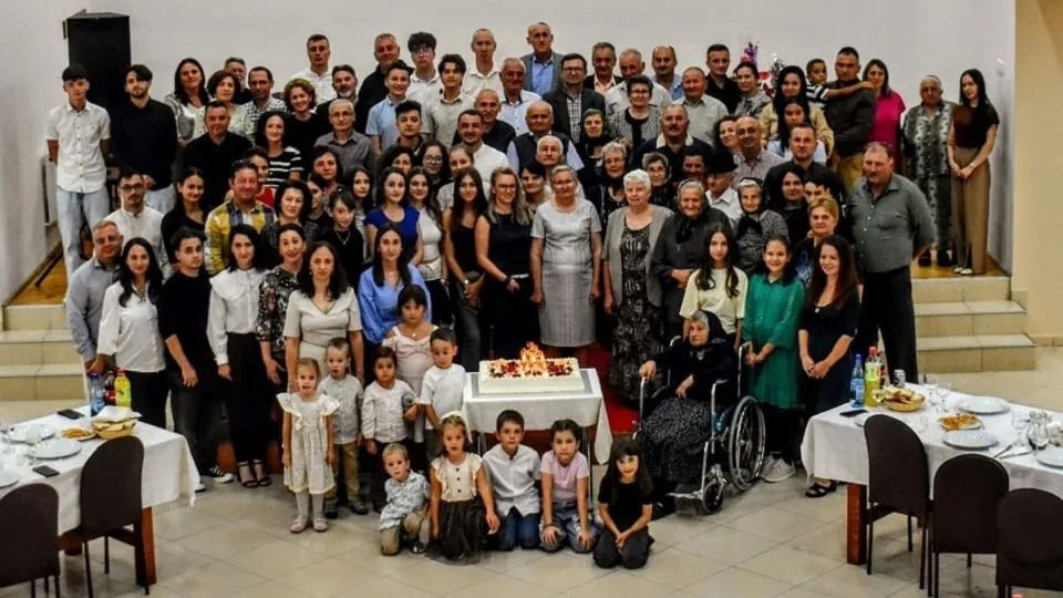 Un grup numeros de adulți și copii pozează în jurul unei femei în scaun rulant, lângă o masă cu tort.