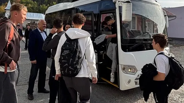 Mai mulți tineri cu rucsacuri stau lângă un autobuz alb cu ușa deschisă, iar câțiva adulți se află în apropiere.