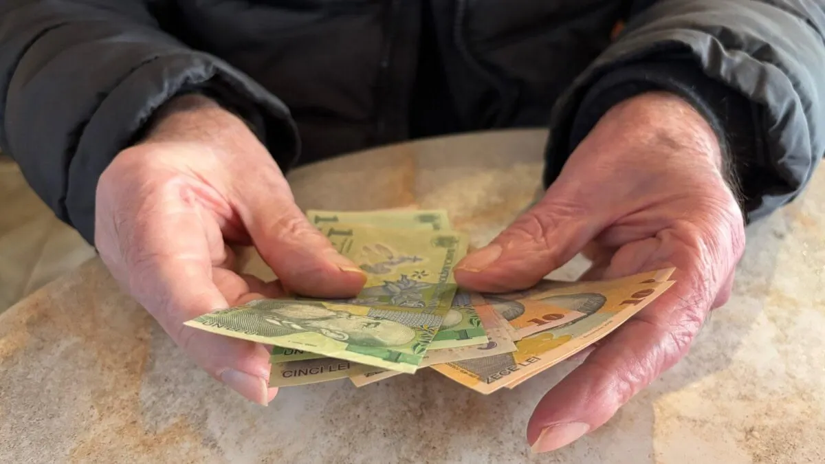 Două mâini cu piele ridată țin mai multe bancnote colorate deasupra unei mese, iar persoana poartă o geacă închisă la culoare.