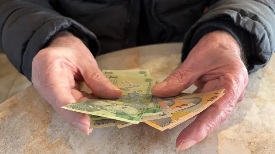 Două mâini cu piele ridată țin mai multe bancnote colorate deasupra unei mese, iar persoana poartă o geacă închisă la culoare.