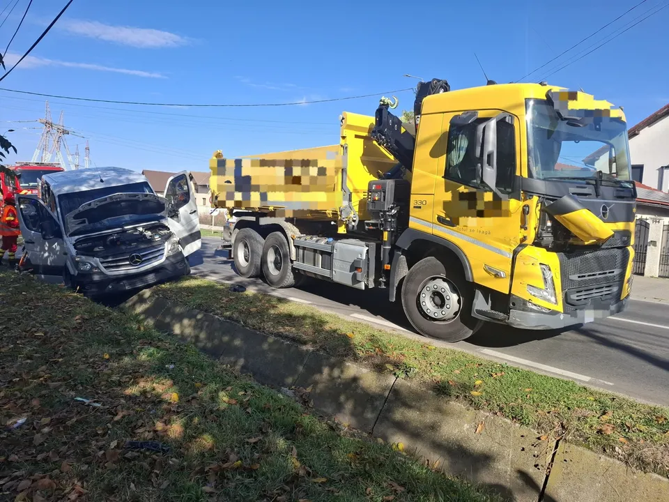 Un autocamion galben se află pe șosea, lângă un microbuz închis la culoare, cu partea din față avariată, înclinat spre marginea drumului.
