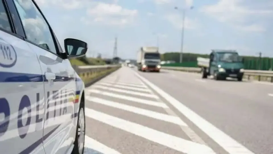 Un autoturism alb cu marcaje albastre ocupă partea stângă, lângă o șosea cu marcaje albe diagonale și mai multe vehicule în depărtare.