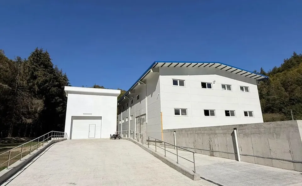 Două clădiri industriale albe se află la capătul unei alei betonate cu balustrade metalice, între versanți împăduriți, sub cer senin.
