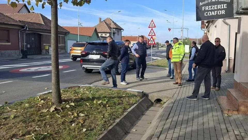 Mai multe persoane stau pe un trotuar lângă un șanț de scurgere; în apropiere se văd o mașină, case și indicatoare rutiere.