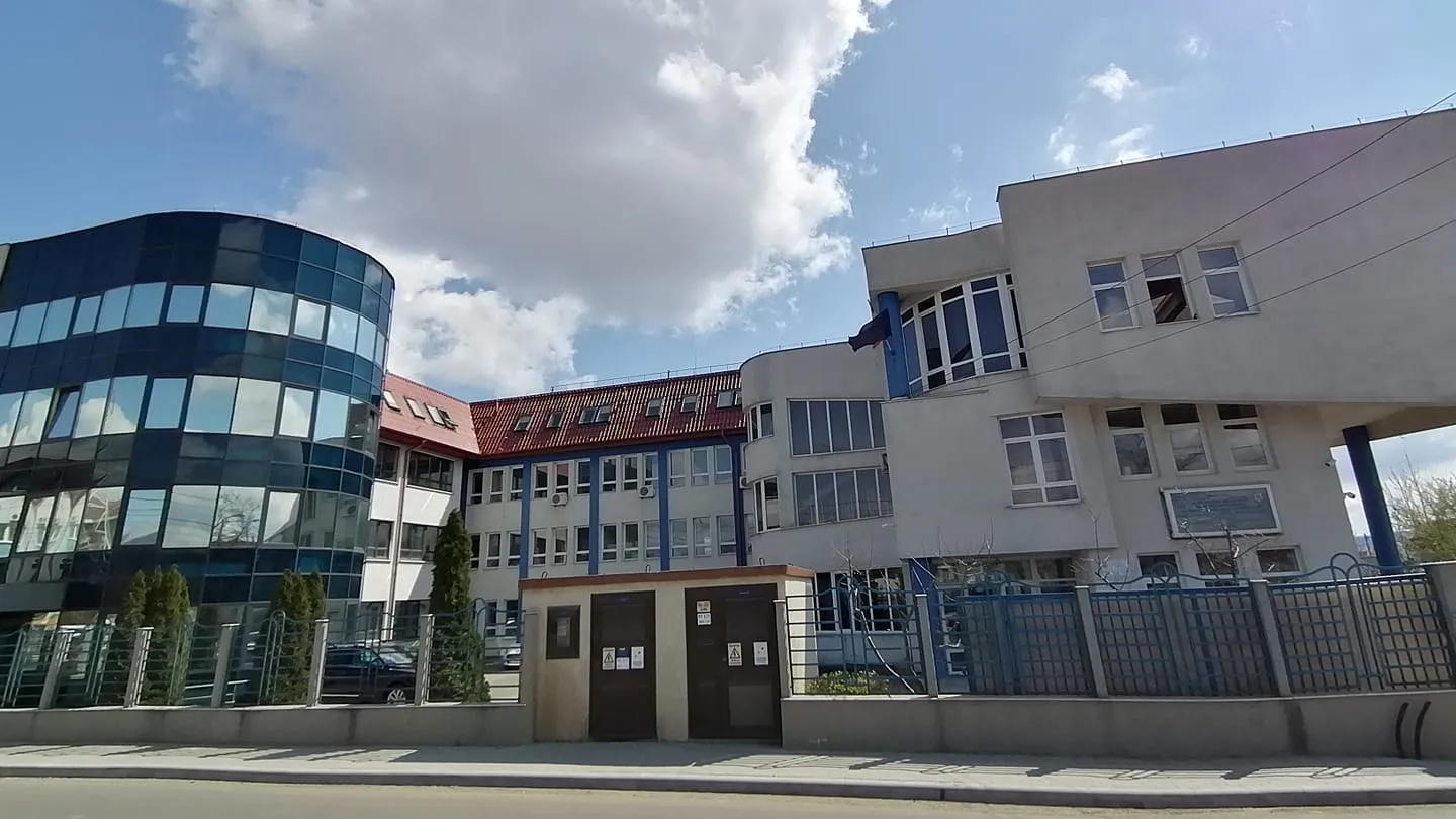 Un ansamblu de clădiri cu fațade vitrate și pereți deschiși la culoare se află în spatele unui gard metalic, lângă un trotuar.