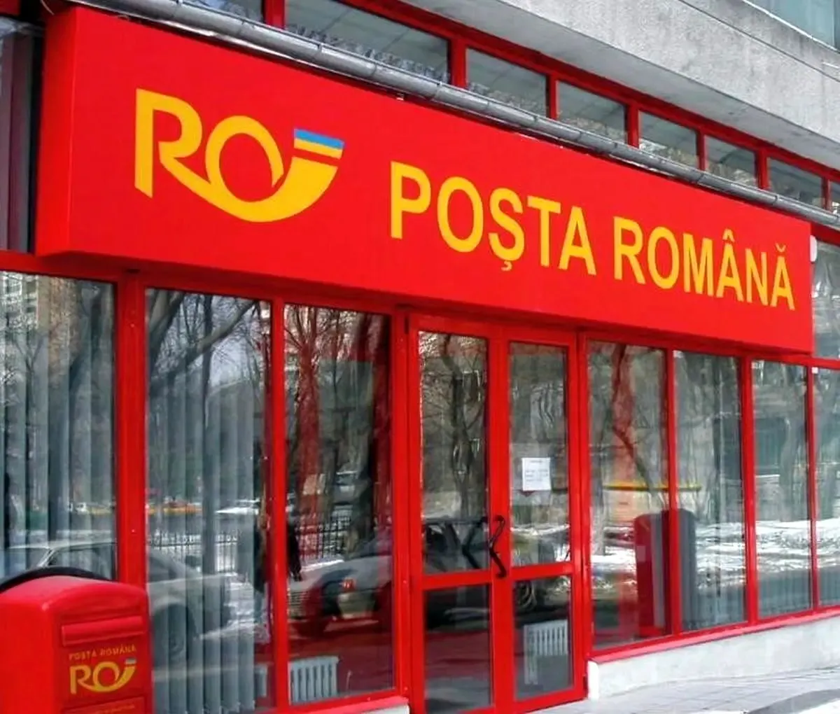 O fațadă cu uși și ferestre de sticlă are deasupra un panou roșu inscripționat „POȘTA ROMÂNĂ”; în stânga se vede o cutie poștală roșie.