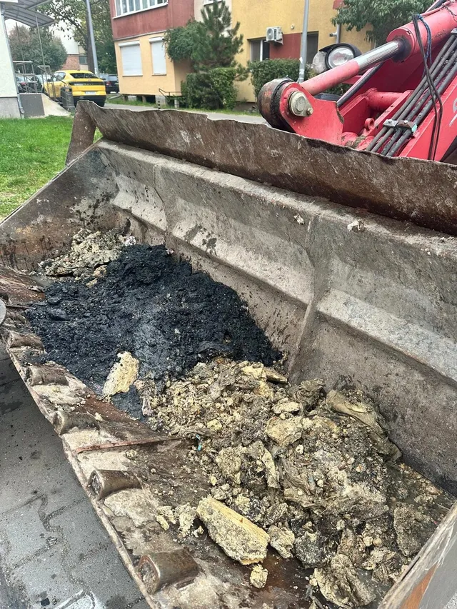 În cupa unui utilaj se află grămezi de material negru și resturi deschise la culoare; în spate se văd brațul roșu al utilajului și o clădire.